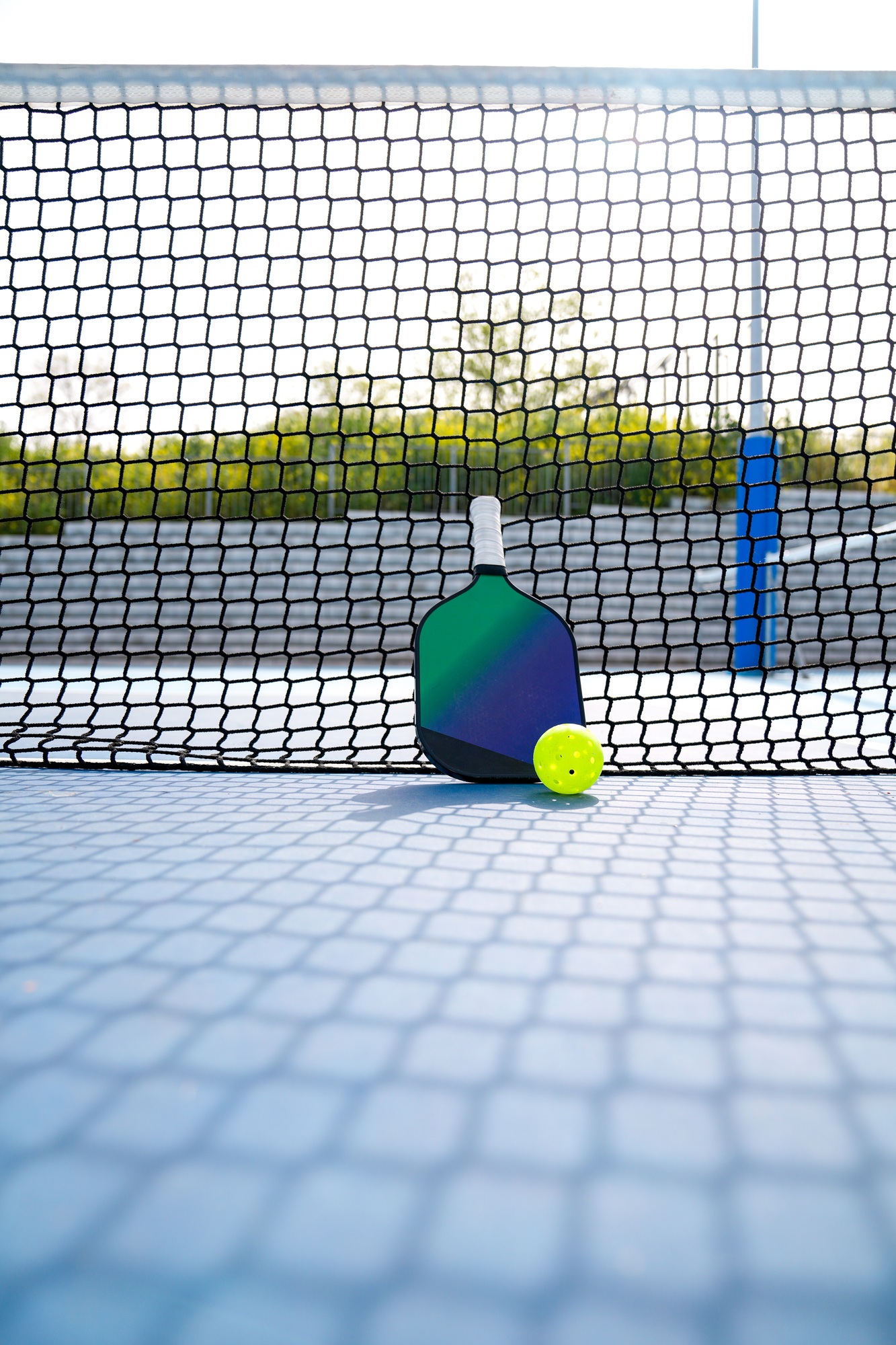 Pickleball paddle and ball leaning against the net on a blue court under sunlight
